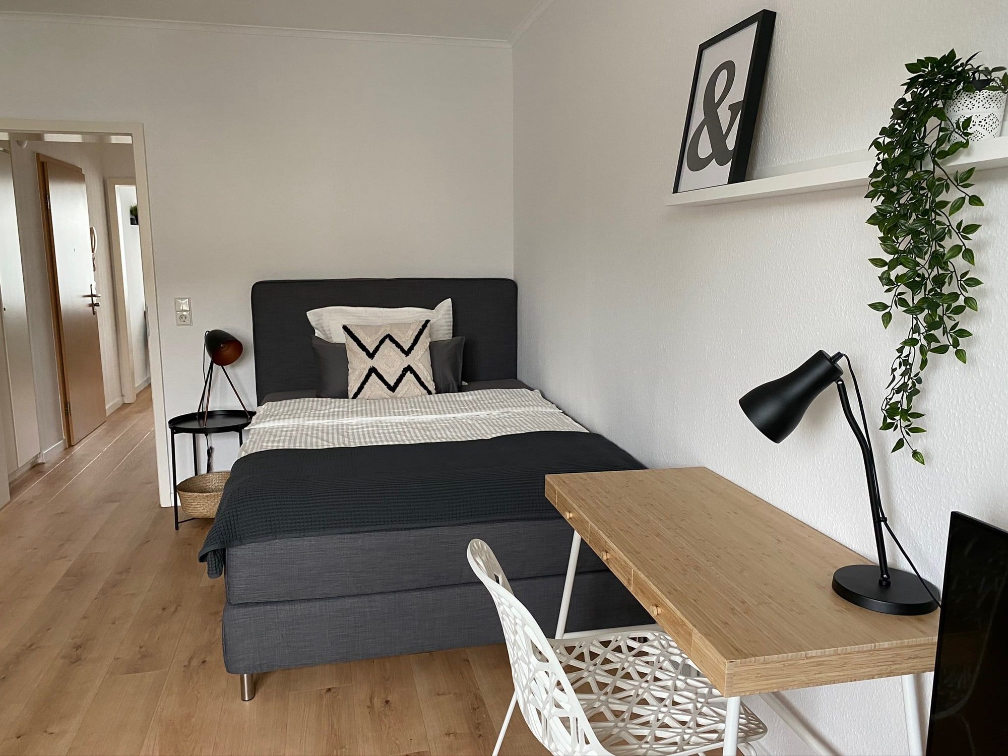 Ein modernes Schlafzimmer mit grauem Bett, Schreibtisch und Pflanze an der Wand.