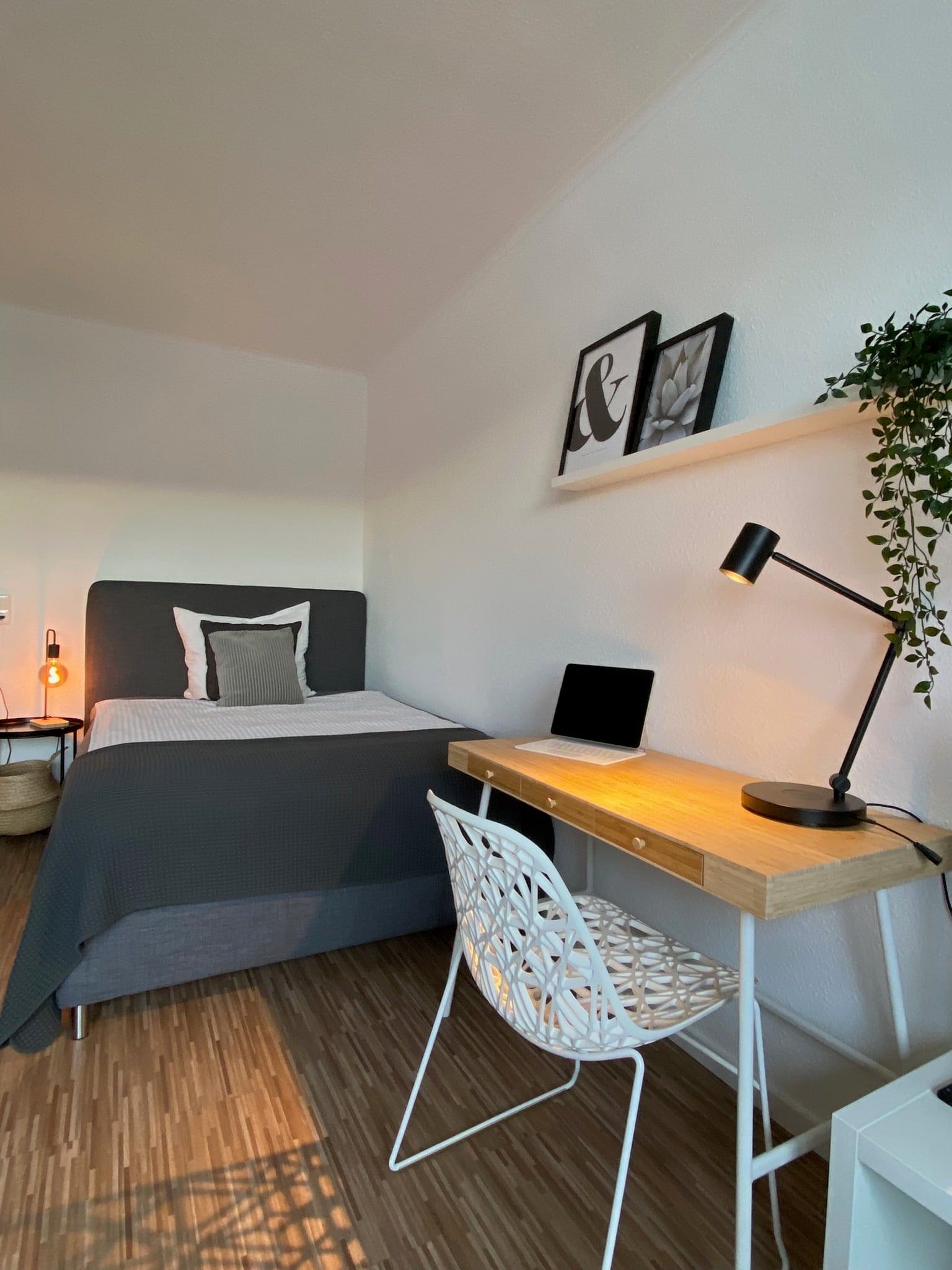A small bedroom with a gray bed and a wooden desk with a lamp and laptop.