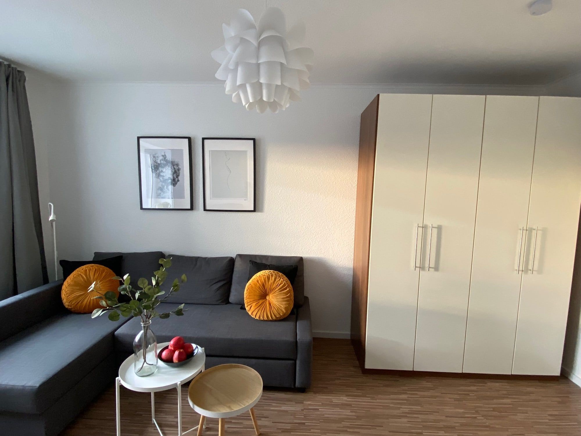 Ein Wohnzimmer mit grauem Ecksofa, zwei orangen Kissen und einem großen weißen Schrank.