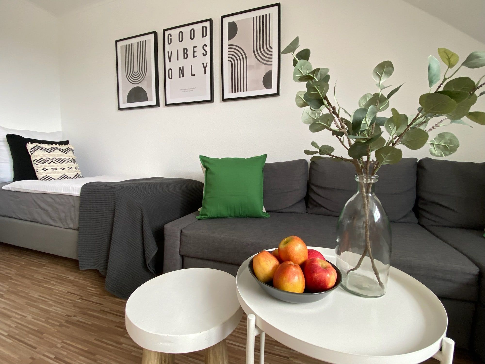 Ein gemütliches Wohnzimmer mit grauem Sofa, grünem Kissen und einem Tisch mit Äpfeln und einer Vase.